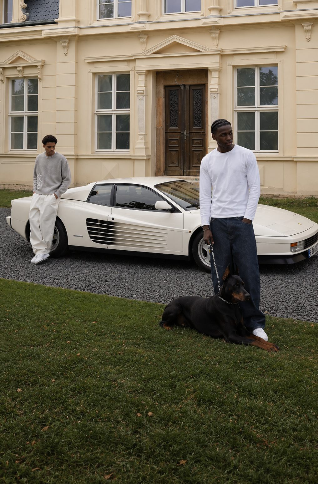 Two men in reda studio pieces beside a white Ferrari Testarossa, a doberman at their feet, in front of a château