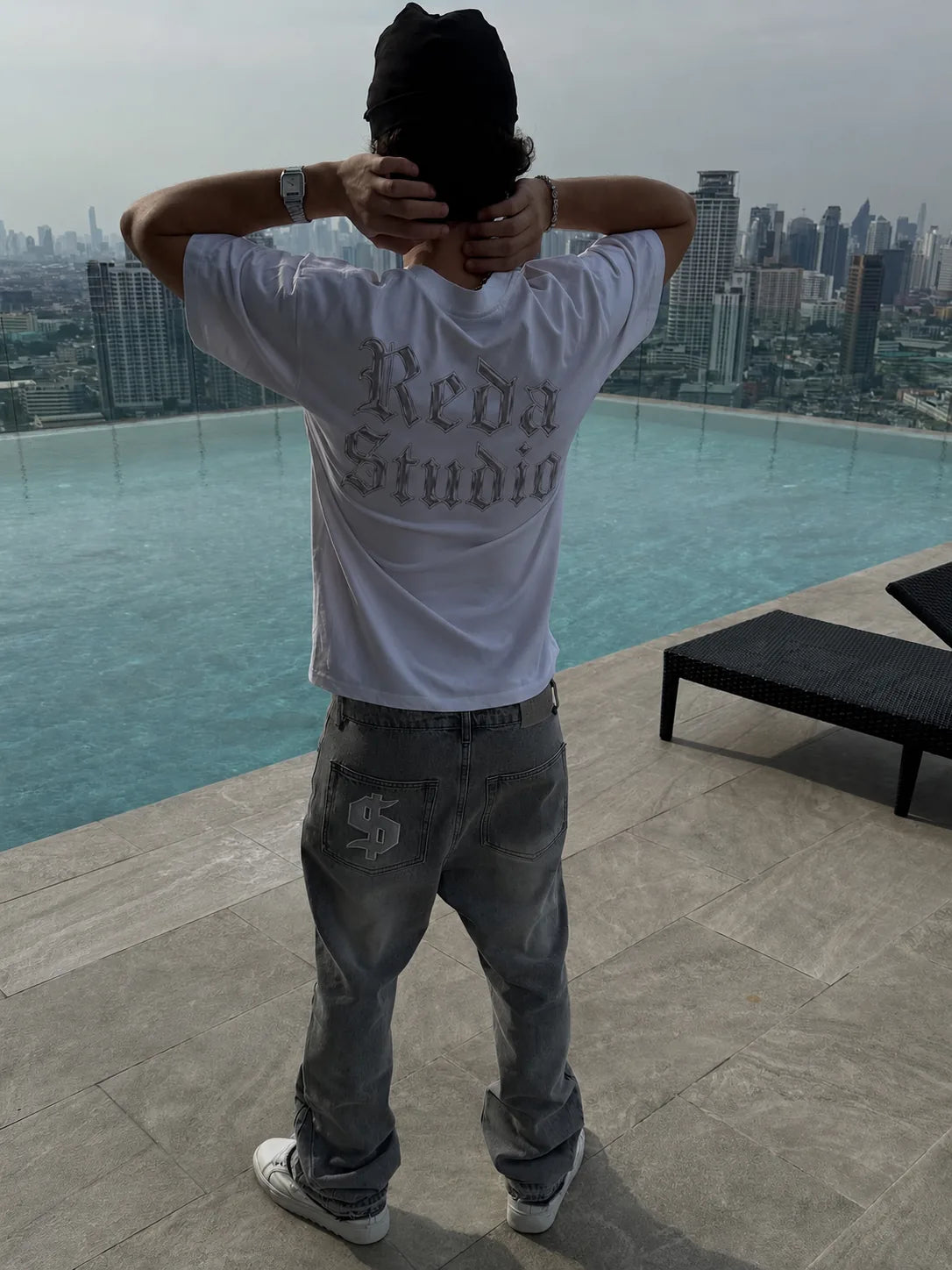 A man from behind in a reda studio t-shirt and jeans, looking out over a rooftop pool and the city skyline