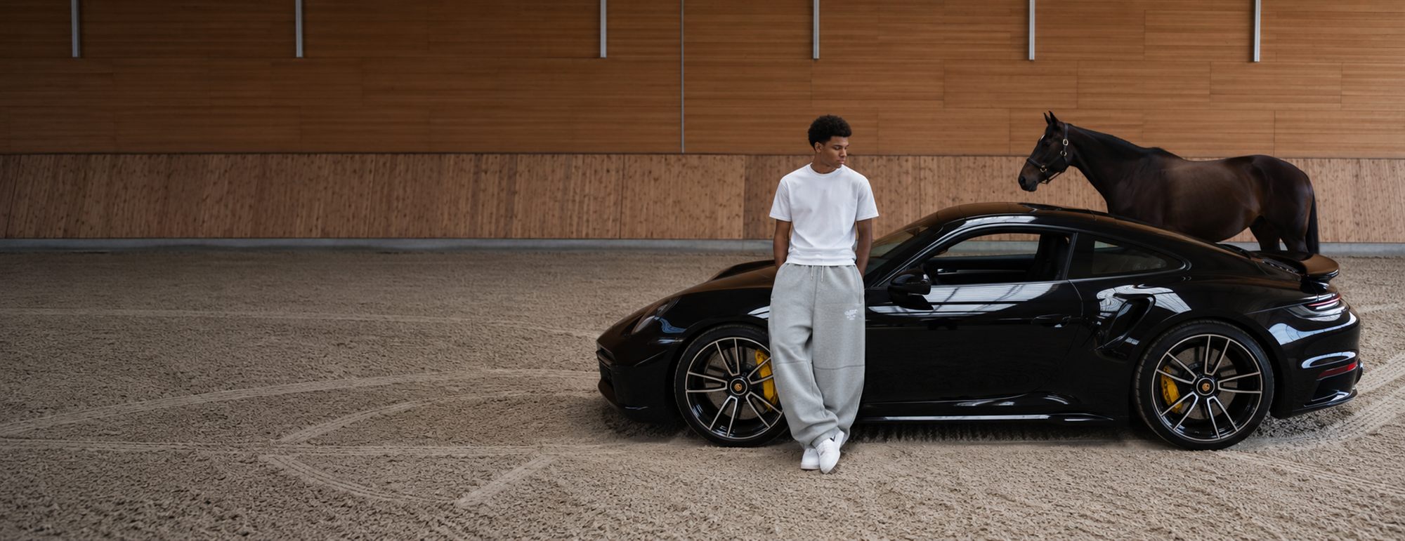 A man in a reda studio t-shirt and sweatpants leaning against a black Porsche, a horse behind him
