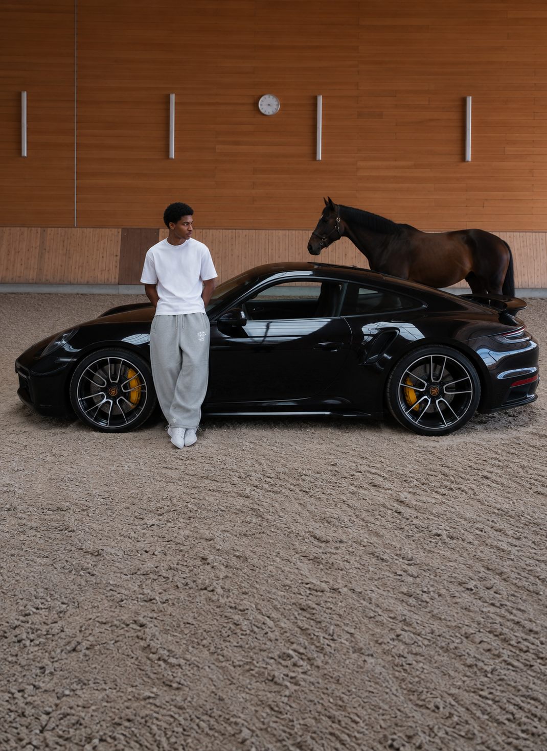 A man in a reda studio t-shirt and sweatpants leaning against a black Porsche, a horse behind him